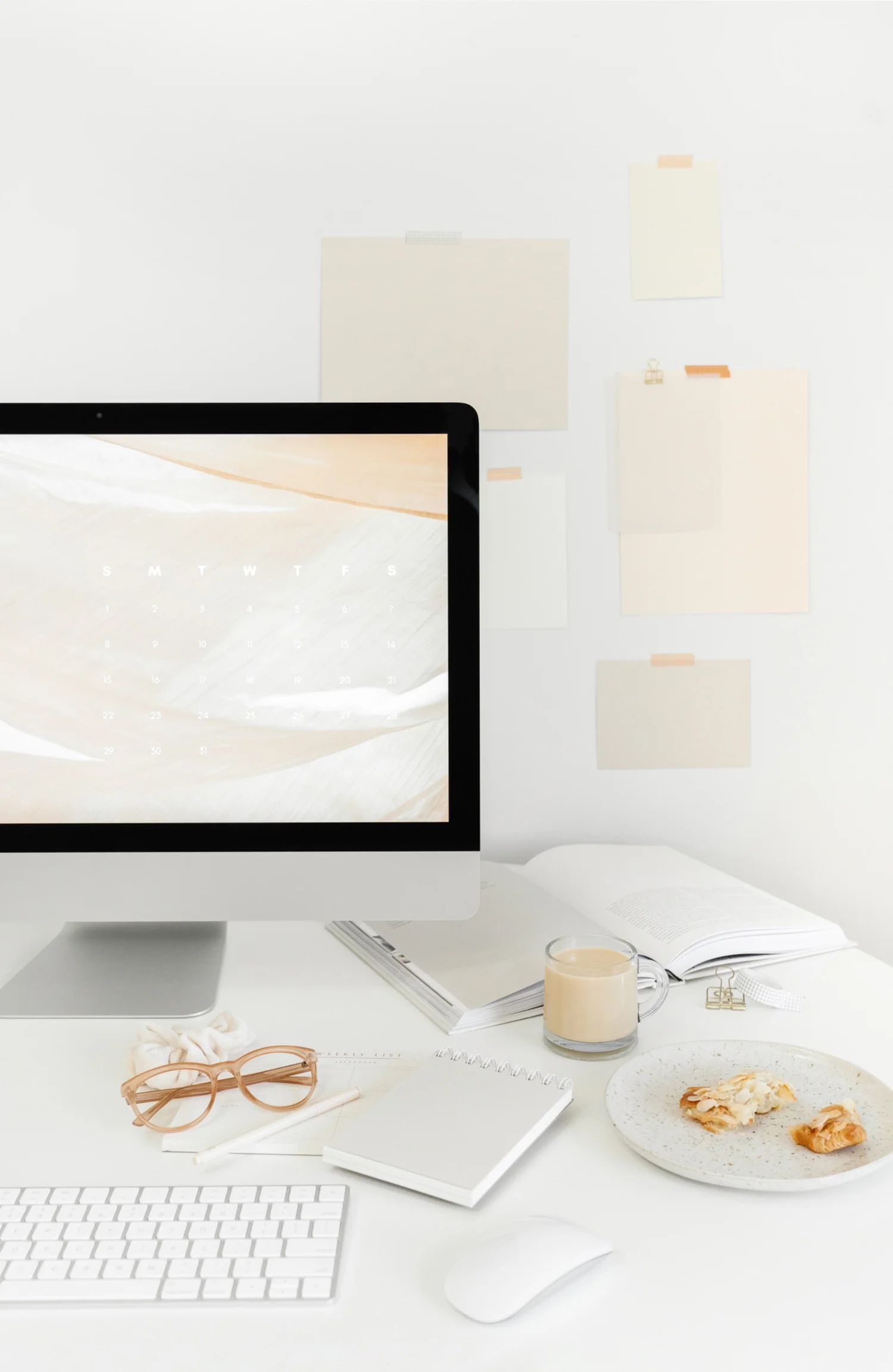 A white workspace with an iMac, notebooks, glasses and croissants