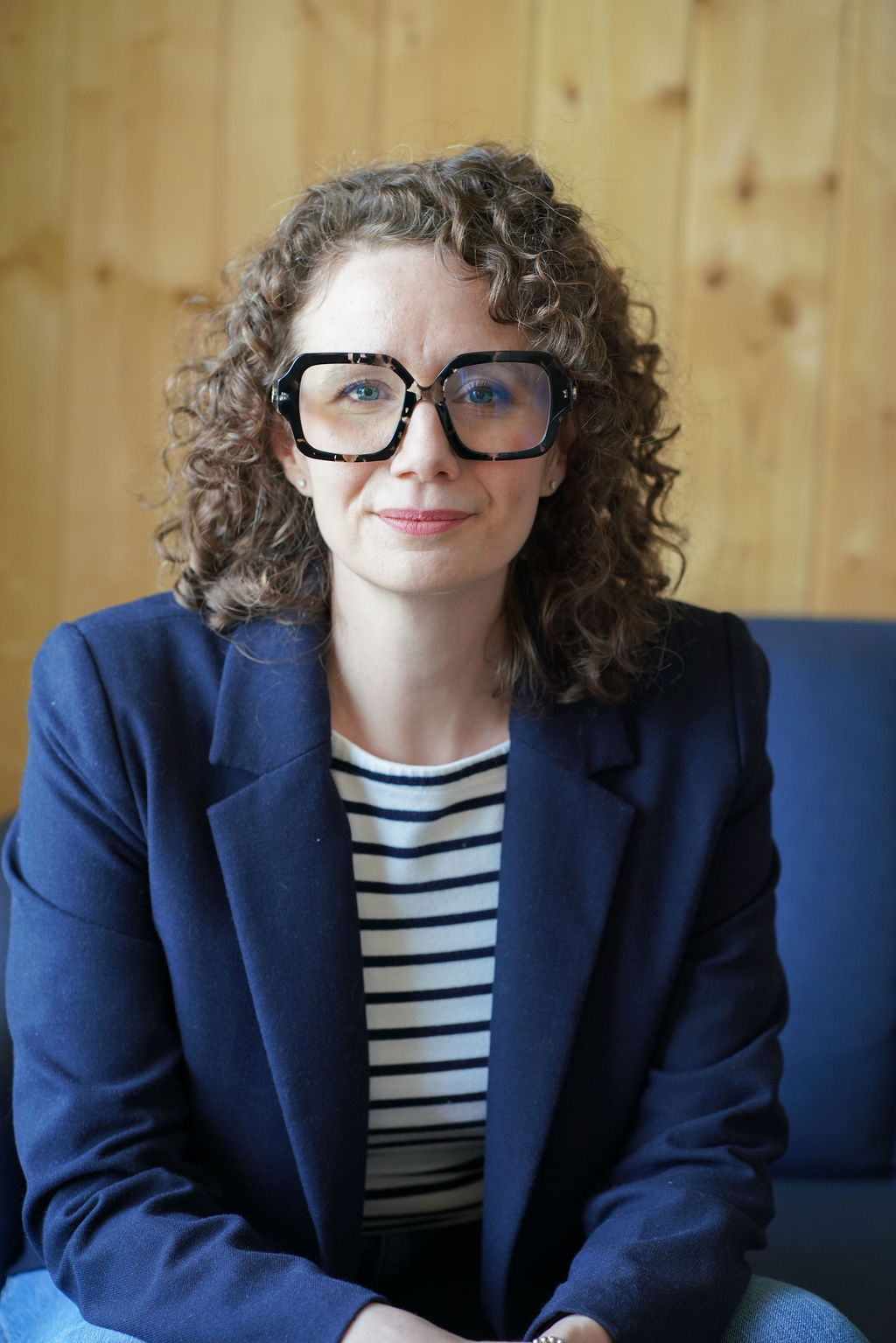 Jessie, with curly hair and large black glasses, in a navy blazer and striped shirt