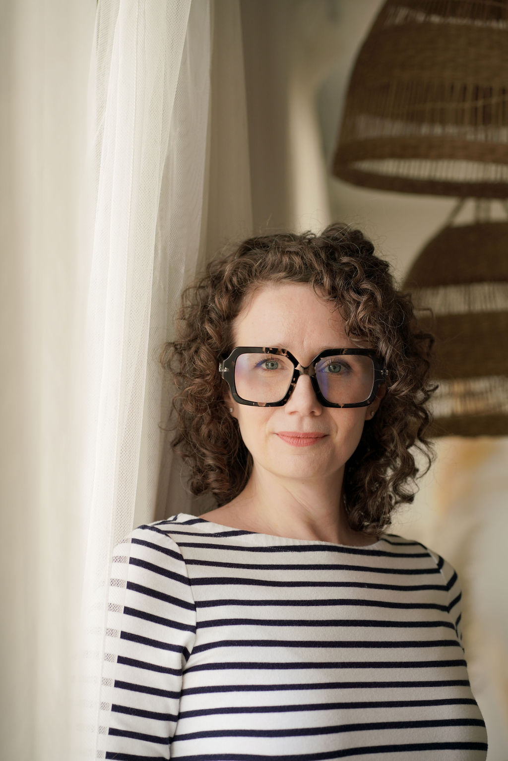 Portrait of Jessie in a striped top beside a sheer curtain, smiling