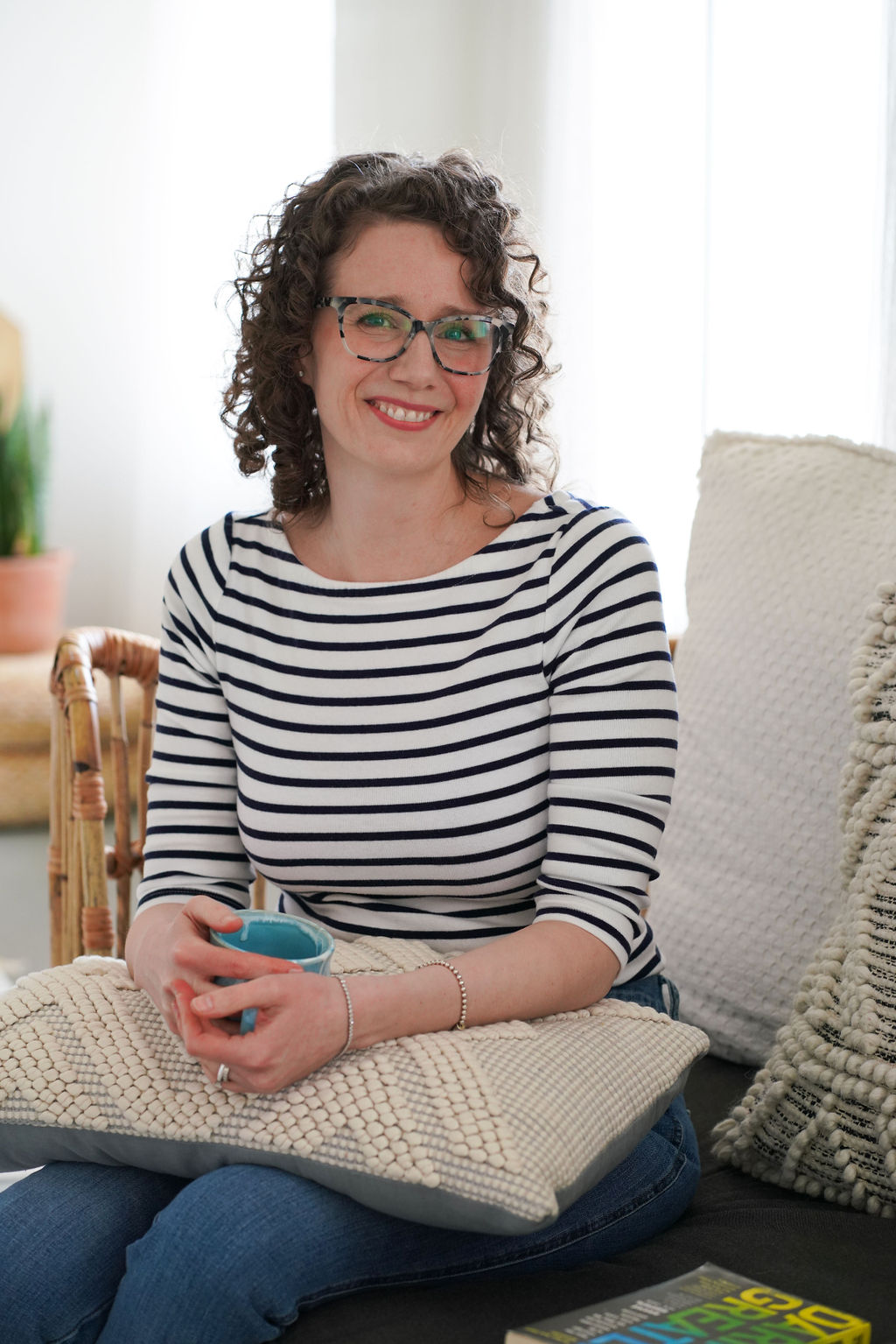 Jessie smiling on a sunlit sofa, holding a blue mug, wearing a striped top